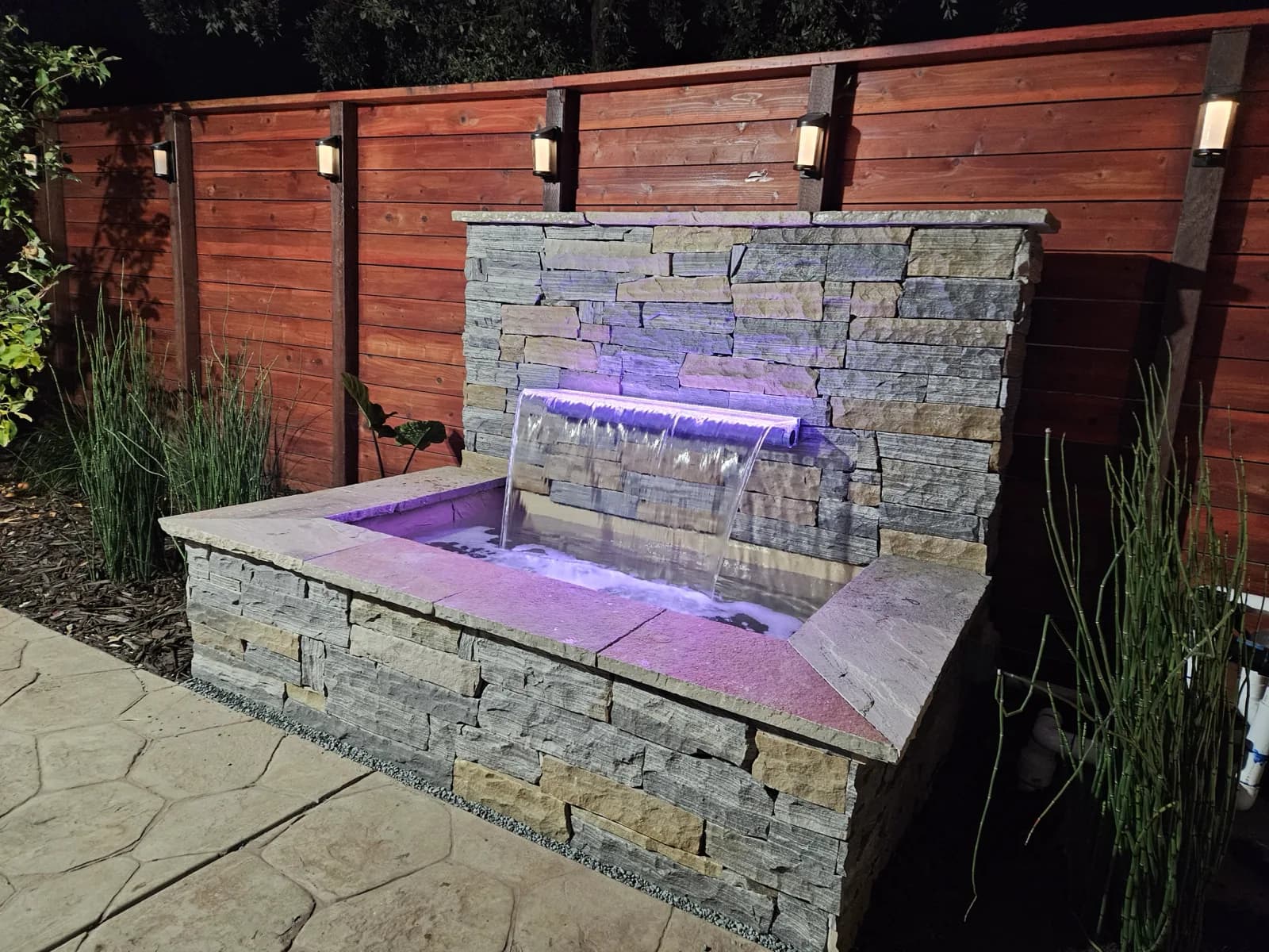 Stacked-stone water feature with a lit spillway in front of a horizontal redwood fence at night