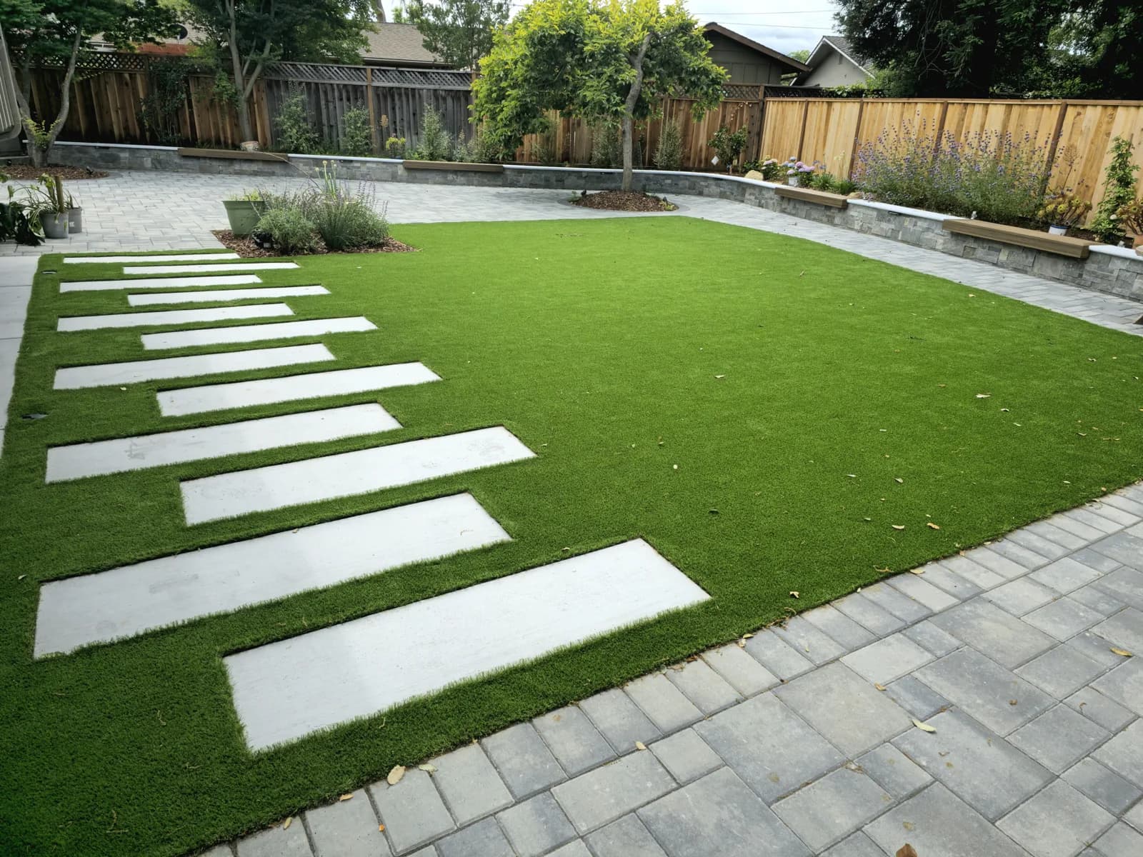 Backyard artificial turf lawn with concrete stepping pads, a paver border and raised planter beds